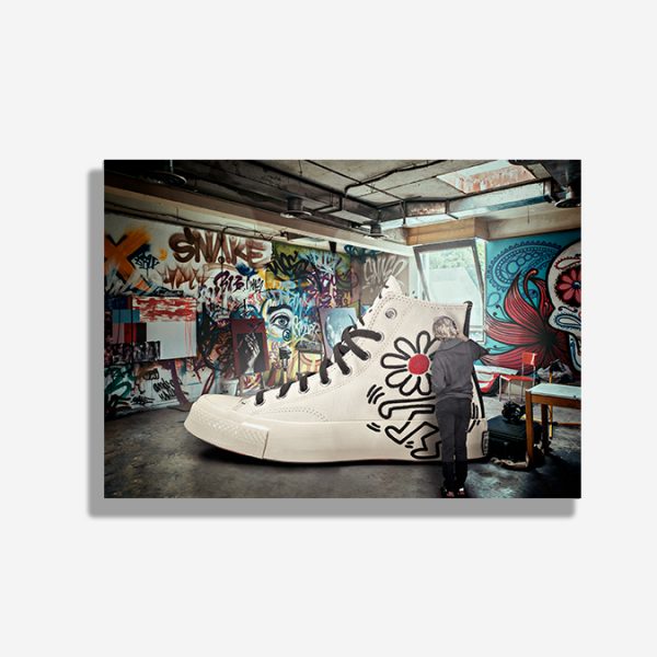 A4 print mockup on a dark background - The image features a Converse Chuck Taylor hi top sneaker with a Keith Haring design. The sneaker is shown as if being painted by an artist in his studio.